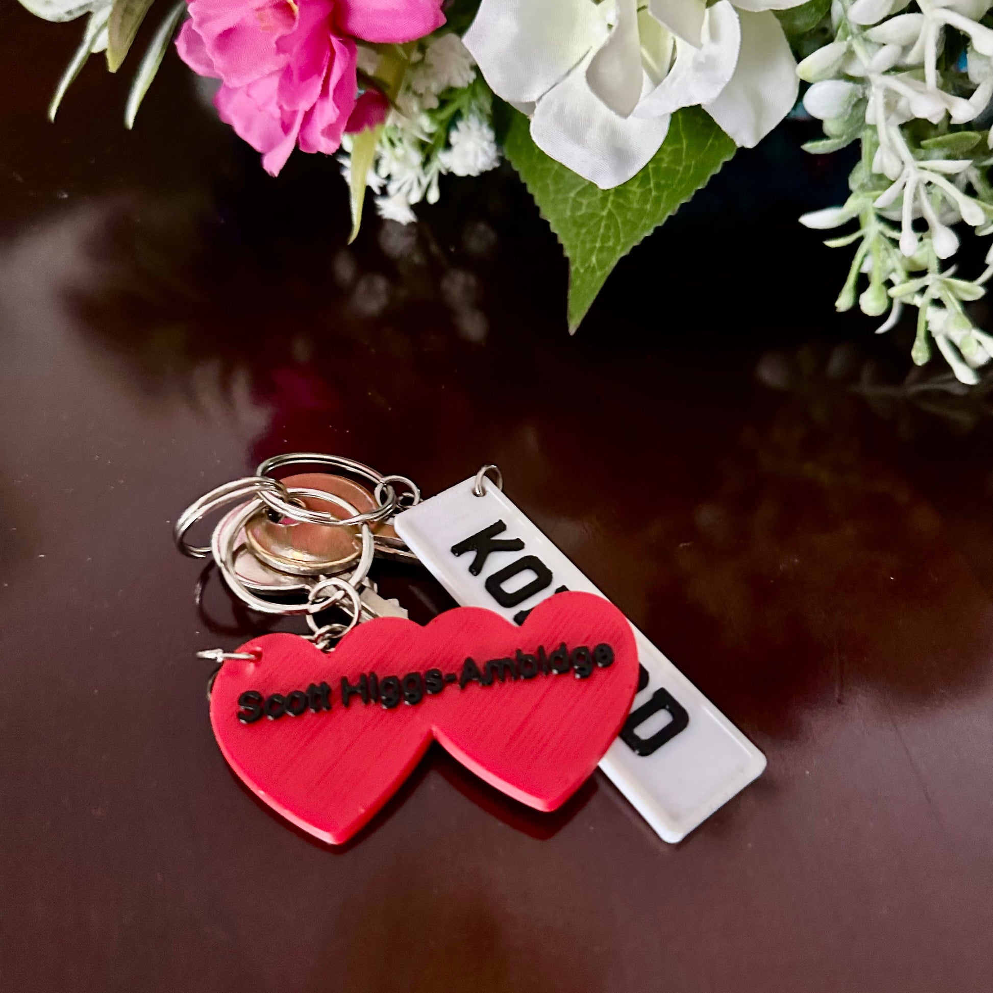 Keychain with red heart and white tag on a reflective surface with flowers in the background