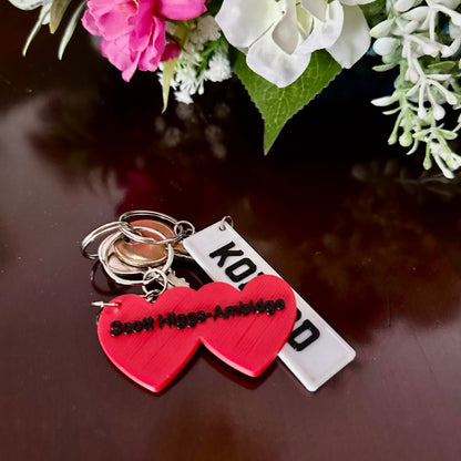 Keychain with red heart and white tag on a reflective surface with flowers in the background