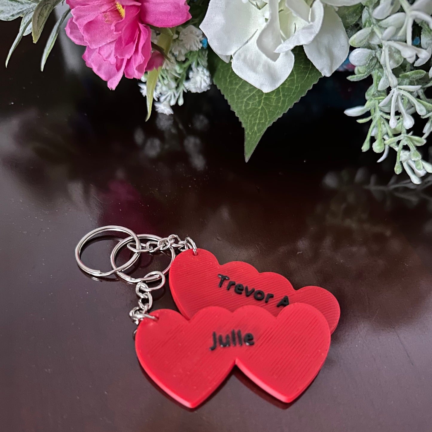 Red heart-shaped keychain with names on a reflective surface with flowers in the background