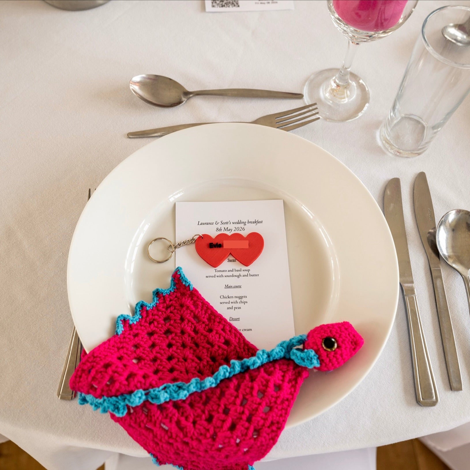 Table setting with a pink and blue crochet dragon, white plate, and silverware on a white tablecloth.
